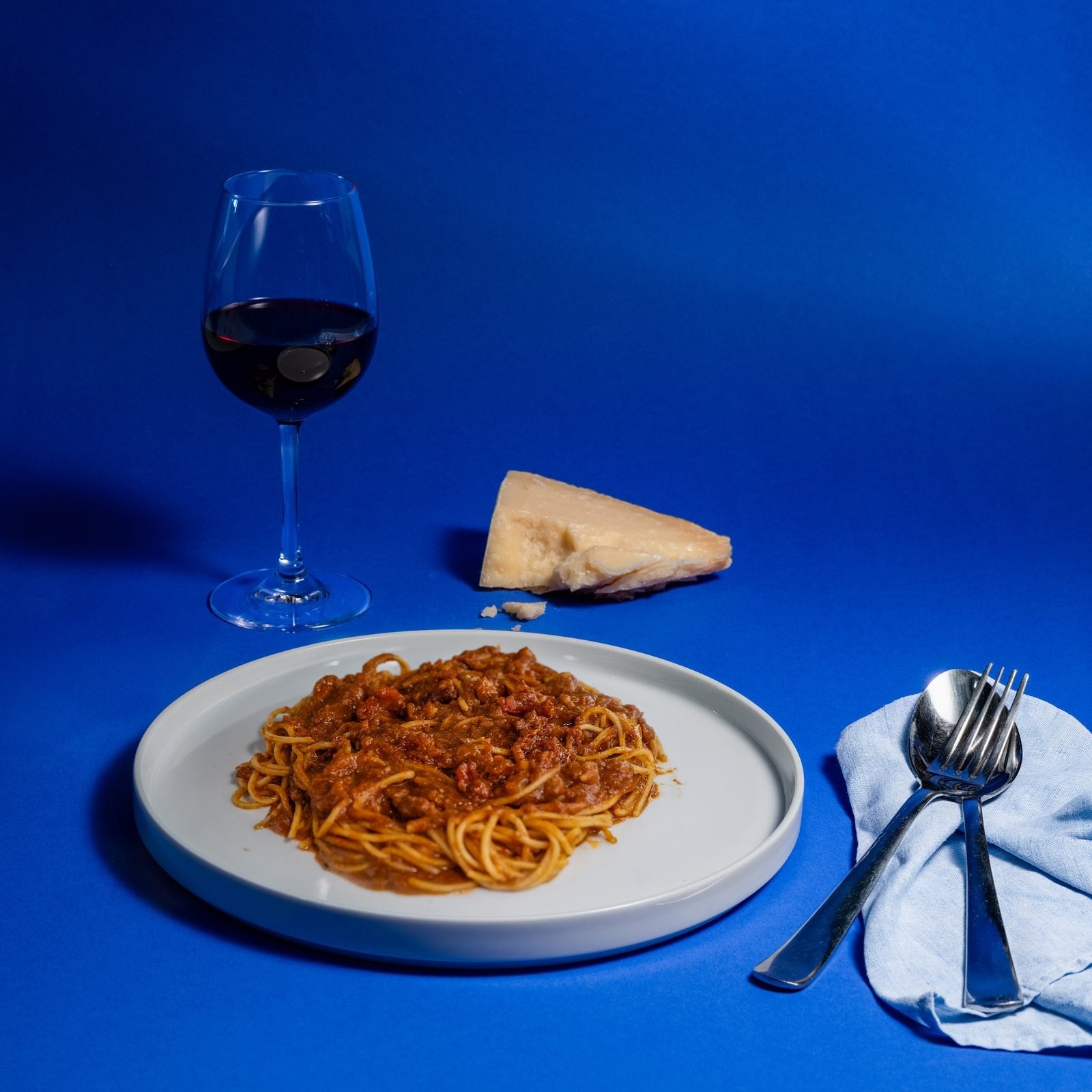 Ein Teller Spaghetti mit Bolognese, ein Glas Rotwein und ein Stück Parmigiano Reggiano auf blauem Studiogrund